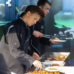 Cynamon Holding Sp. z o.o. - Dwóch kucharzy w czarnych uniformach przygotowuje dania z grilla na stanowisku cateringowym, jeden nakłada szaszłyki warzywne na talerz, drugi podaje gotowe mięso.
