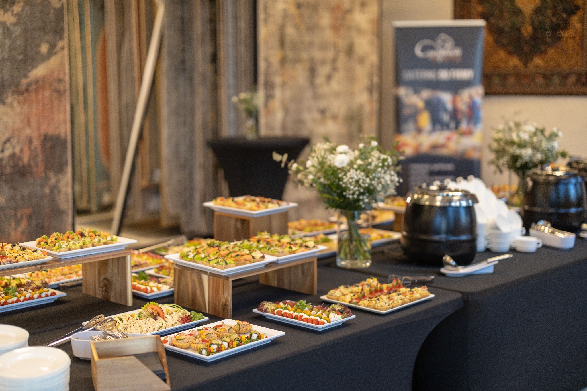 Stół cateringowy z przekąskami na białych talerzach, drewniane podesty, kwiaty w wazonie, podgrzewacze do potraw, czarny obrus, w tle dekoracyjne zasłony i baner reklamowy.