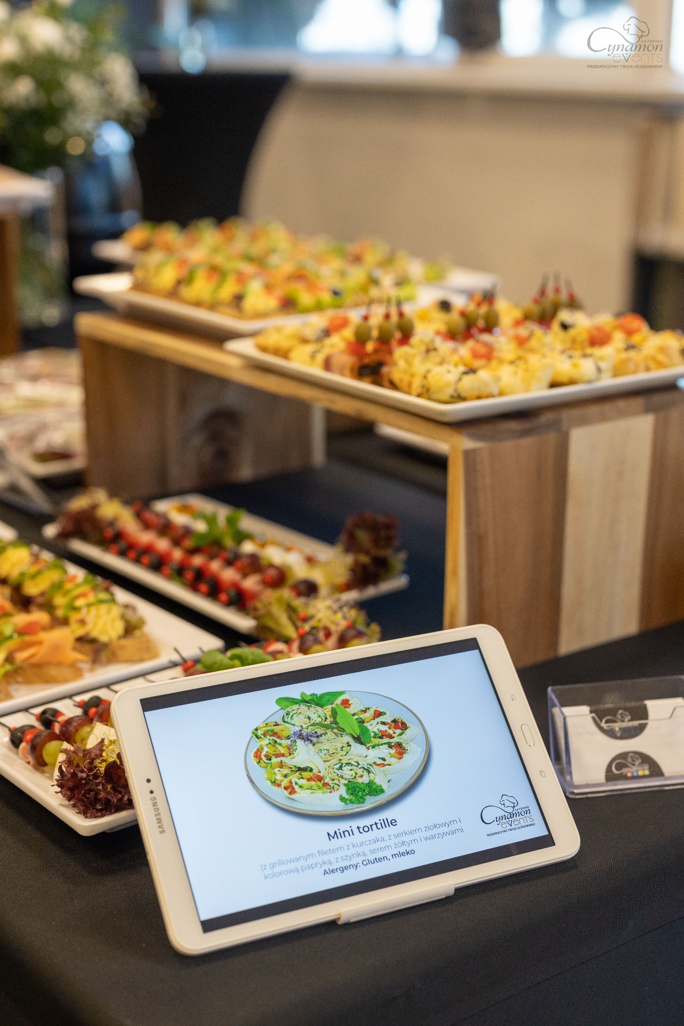 Stół cateringowy z przekąskami na białych talerzach, w tle kwiaty, na pierwszym planie tablet wyświetlający zdjęcie mini tortilli z opisem składników i logo firmy cateringowej 'Cynamon Events'...
