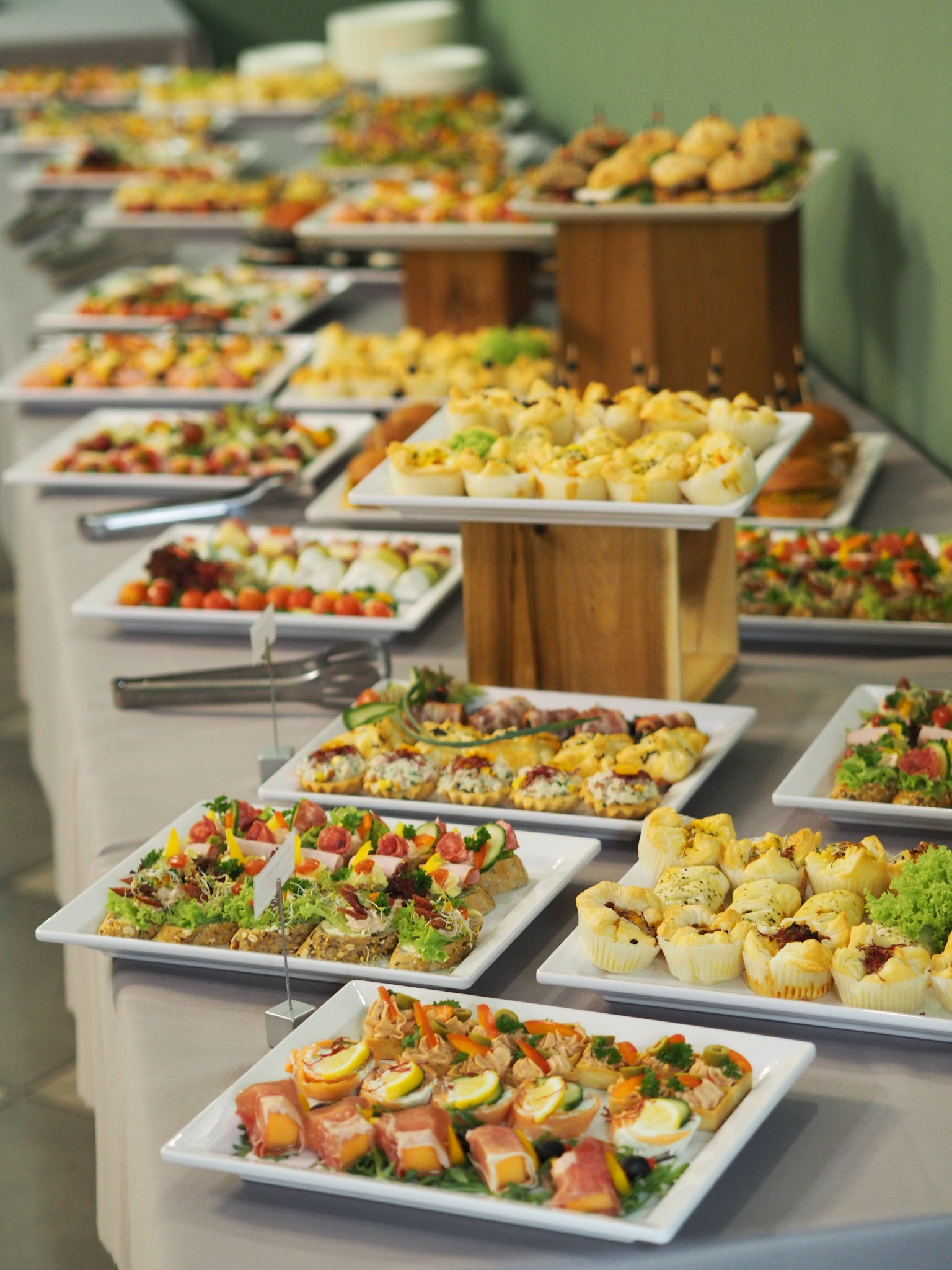 Długi stół nakryty szarym obrusem, zastawiony białymi kwadratowymi talerzami z różnorodnymi przekąskami: mini kanapeczki, tartaletki, roladki z szynki parmeńskiej i melona, ustawione na różnych...