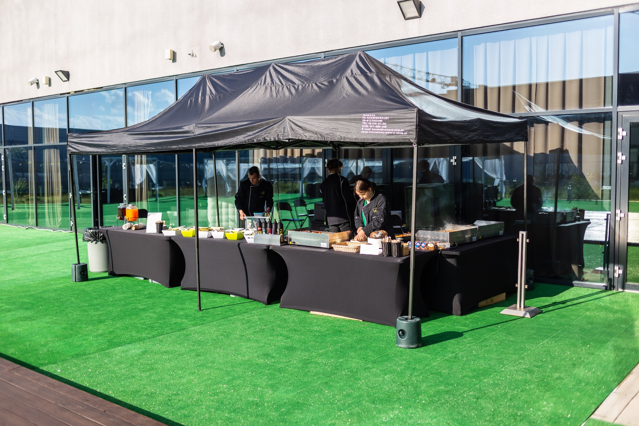 Zewnętrzny punkt gastronomiczny pod czarnym namiotem z widocznym logo firmy, obsługa w strojach roboczych przygotowuje dania na grillu, na stole pojemniki z jedzeniem i napojami, zielona sztuczna...