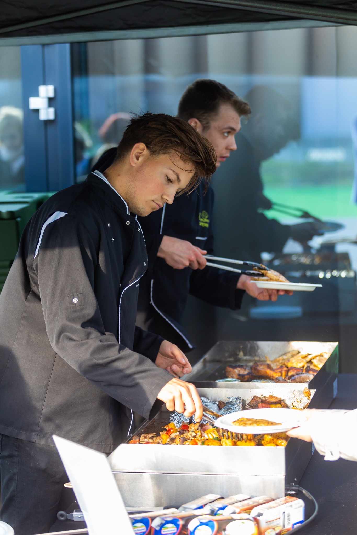 Dwóch kucharzy w czarnych uniformach przygotowuje dania z grilla na stanowisku cateringowym, jeden nakłada szaszłyki warzywne na talerz, drugi podaje gotowe mięso.