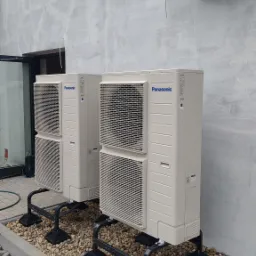 Dwie jednostki zewnętrzne pompy ciepła Panasonic stojące na czarnych, metalowych wspornikach na tle szarej ściany z widoczną jednostką klimatyzacji Kaisai Eco zamontowaną powyżej, obok widoczny...
