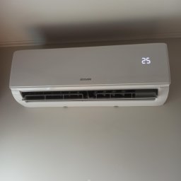 STELAR Przedsiębiorstwo Handlowo - Usługowe Greta Turska - Biała klimatyzacja Sinclair na ścianie, wyświetlająca temperaturę 25 stopni. Widok z dołu, oświetlenie naturalne.