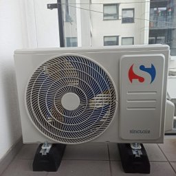 STELAR Przedsiębiorstwo Handlowo - Usługowe Greta Turska - Kompaktowa jednostka zewnętrzna klimatyzacji Sinclair na balkonie, zamontowana na czarnych podkładkach antywibracyjnych. Widok na budynek przez szklaną balustradę.
