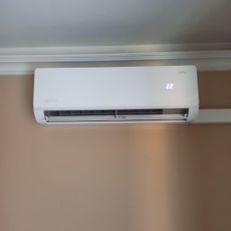 STELAR Przedsiębiorstwo Handlowo - Usługowe Greta Turska - Biały klimatyzator ścienny marki Roni z wyświetlaczem temperatury 22°C, zamontowany w pomieszczeniu o beżowych ścianach i białym suficie z dekoracyjną listwą.