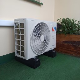 STELAR Przedsiębiorstwo Handlowo - Usługowe Greta Turska - Biała jednostka zewnętrzna klimatyzatora Sinclair na czarnych podkładkach antywibracyjnych, stojąca na zielonej sztucznej trawie. W tle żółta ściana z oknem i drewniana donica z roślinami.