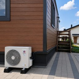 STELAR Przedsiębiorstwo Handlowo - Usługowe Greta Turska - Zewnętrzna jednostka klimatyzacji GREE na czarnych podstawach, zainstalowana przy nowoczesnym domu z brązową elewacją i ciemnymi oknami. Widoczne drewniane schody i kostka brukowa.