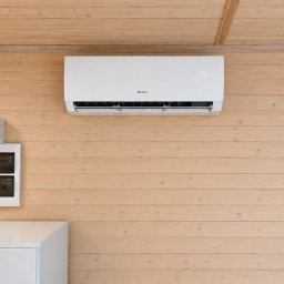 STELAR Przedsiębiorstwo Handlowo - Usługowe Greta Turska - Biały klimatyzator GREE z wyświetlaczem '23' zamontowany na jasnej, drewnianej ścianie z poziomych desek, obok nowoczesna rozdzielnica elektryczna i biała szafka.