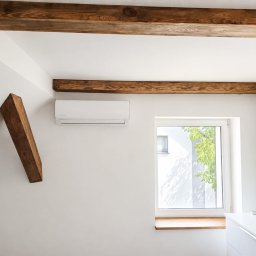 STELAR Przedsiębiorstwo Handlowo - Usługowe Greta Turska - Biała klimatyzacja ścienna na tle białej ściany z surowymi, drewnianymi belkami stropowymi i ukośną podporą. Obok okno z drewnianym parapetem i zielenią za szybą.