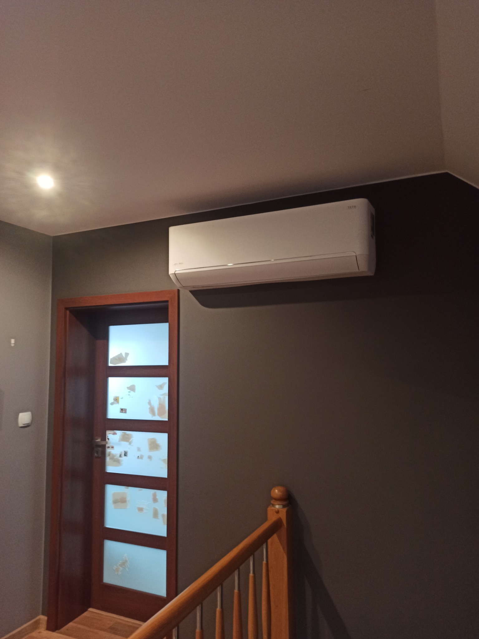 Biały klimatyzator ścienny zamontowany nad drewnianą, przeszkloną drzwiach w szarej klatce schodowej z drewnianą balustradą.
