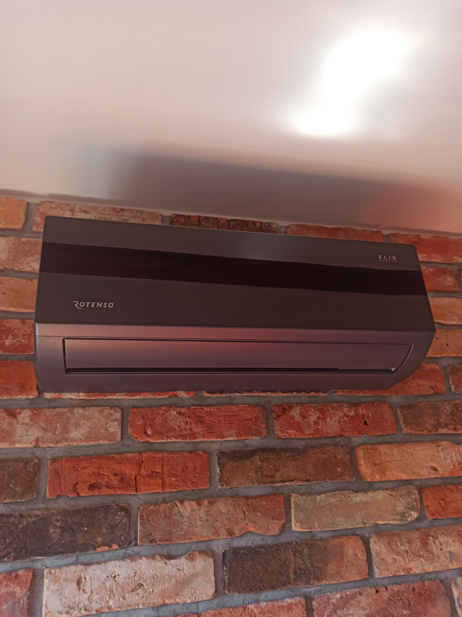 Szary klimatyzator Rotenso Elis Series zamontowany na ścianie z czerwonej cegły w pomieszczeniu z jasnym sufitem.