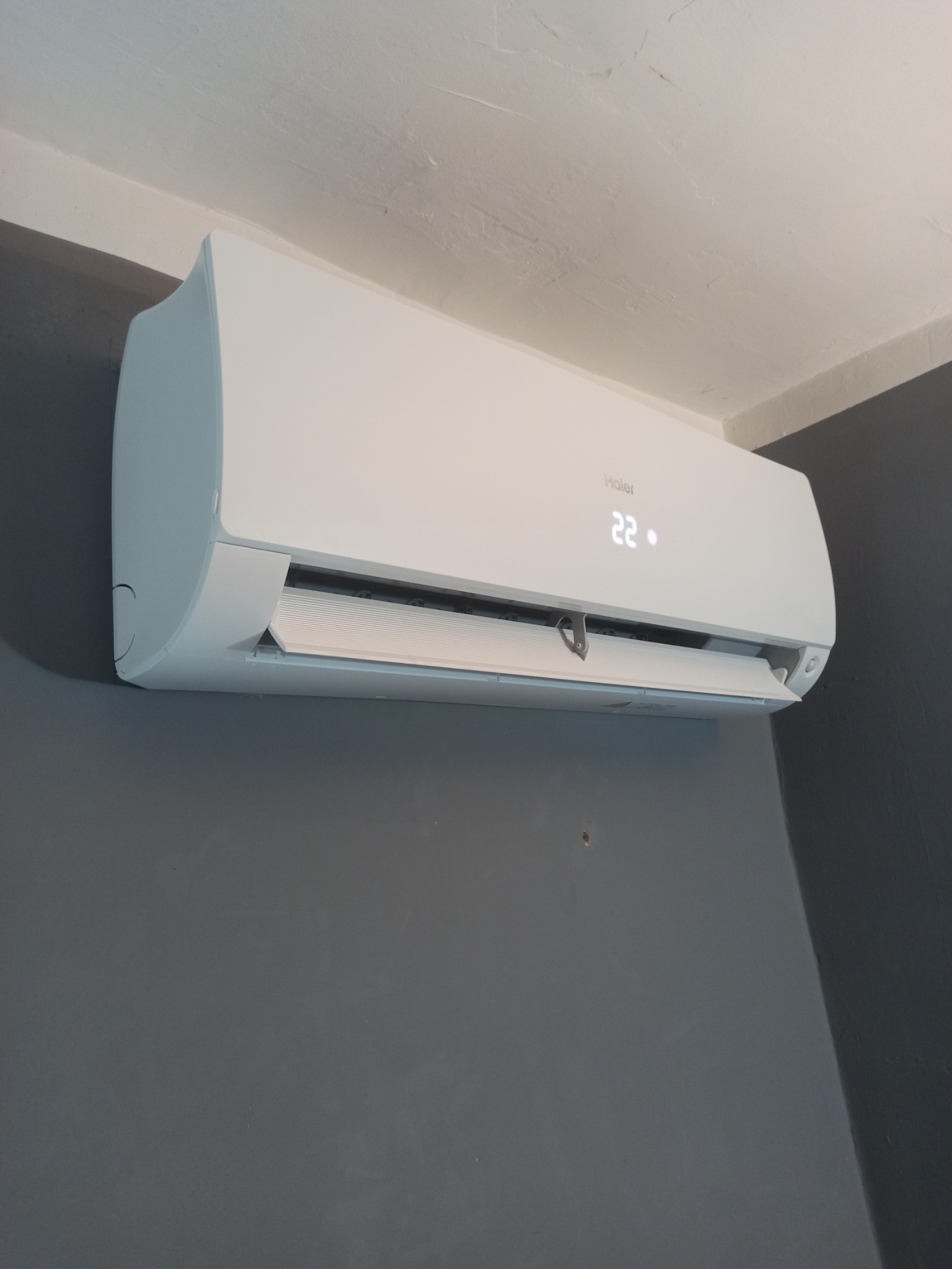 Biała jednostka klimatyzacyjna Haier zamontowana na szarej ścianie, wyświetlająca temperaturę 22 stopnie Celsjusza.