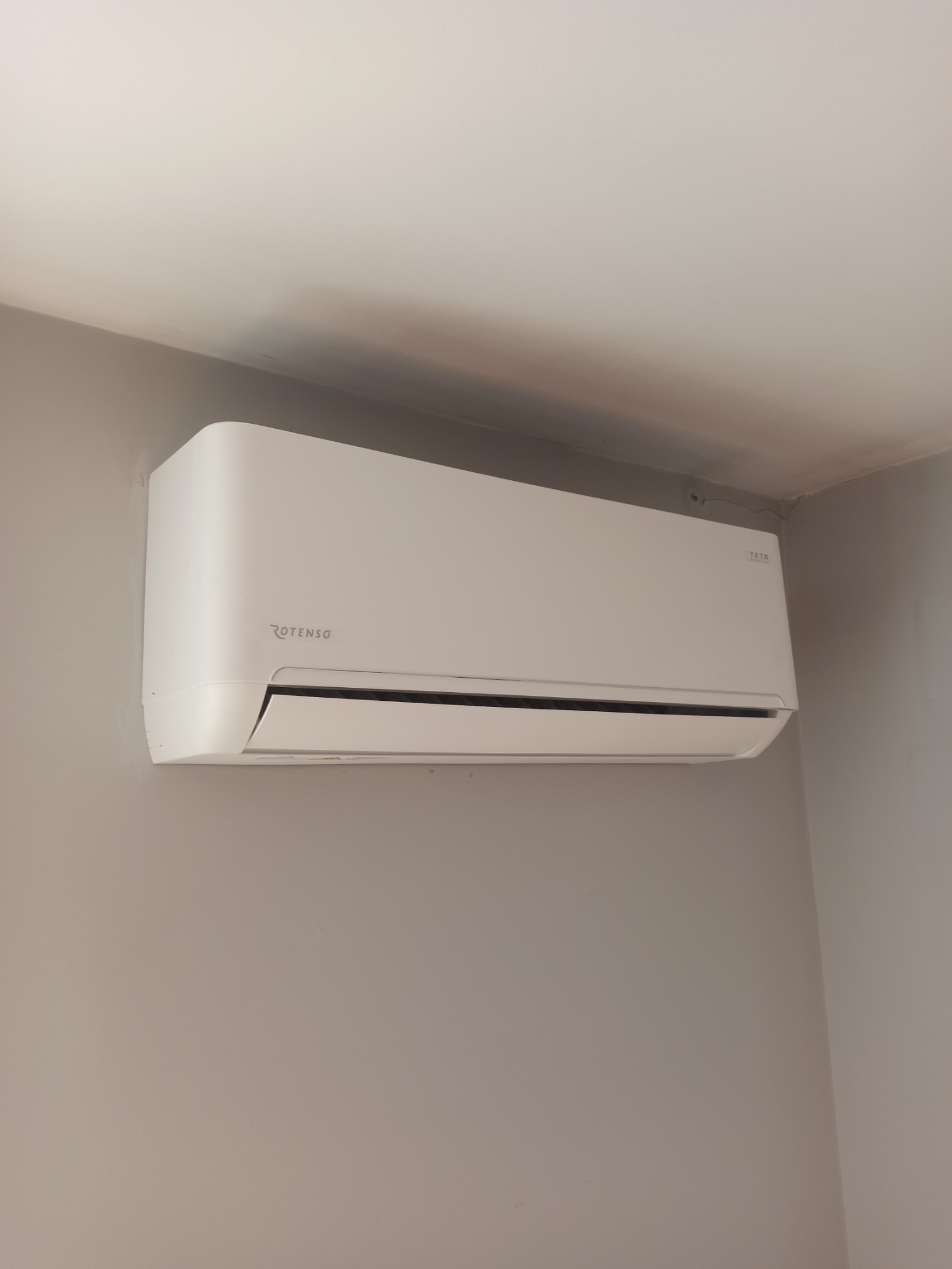 Biała jednostka wewnętrzna klimatyzacji marki Rotenso zamontowana na szarej ścianie w pomieszczeniu.