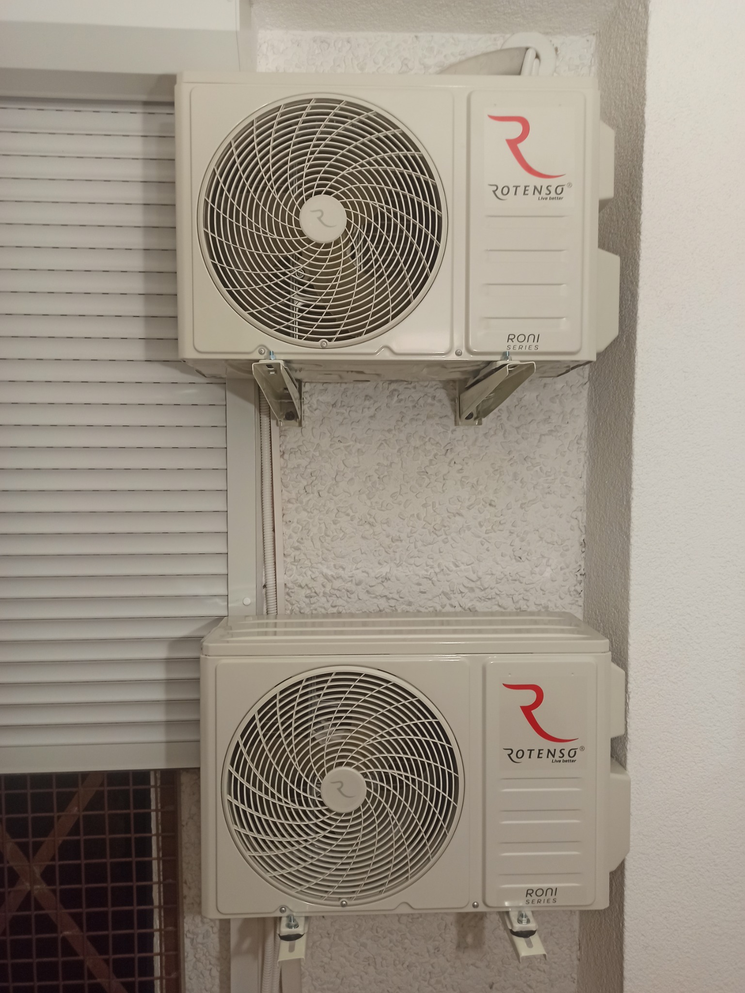 Dwa klimatyzatory marki Rotenso zamontowane jeden nad drugim na ścianie o nierównej fakturze, widoczne metalowe wsporniki mocujące i fragment rolety okiennej.