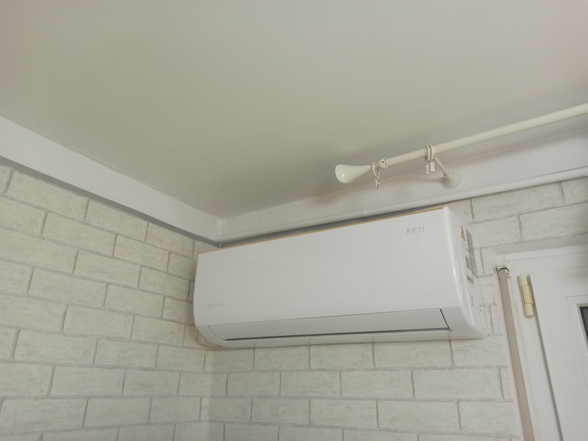 Biały klimatyzator marki Rotenso zamontowany na ścianie z dekoracyjnej cegły, widoczny fragment okna i karnisz z reflektorkiem.
