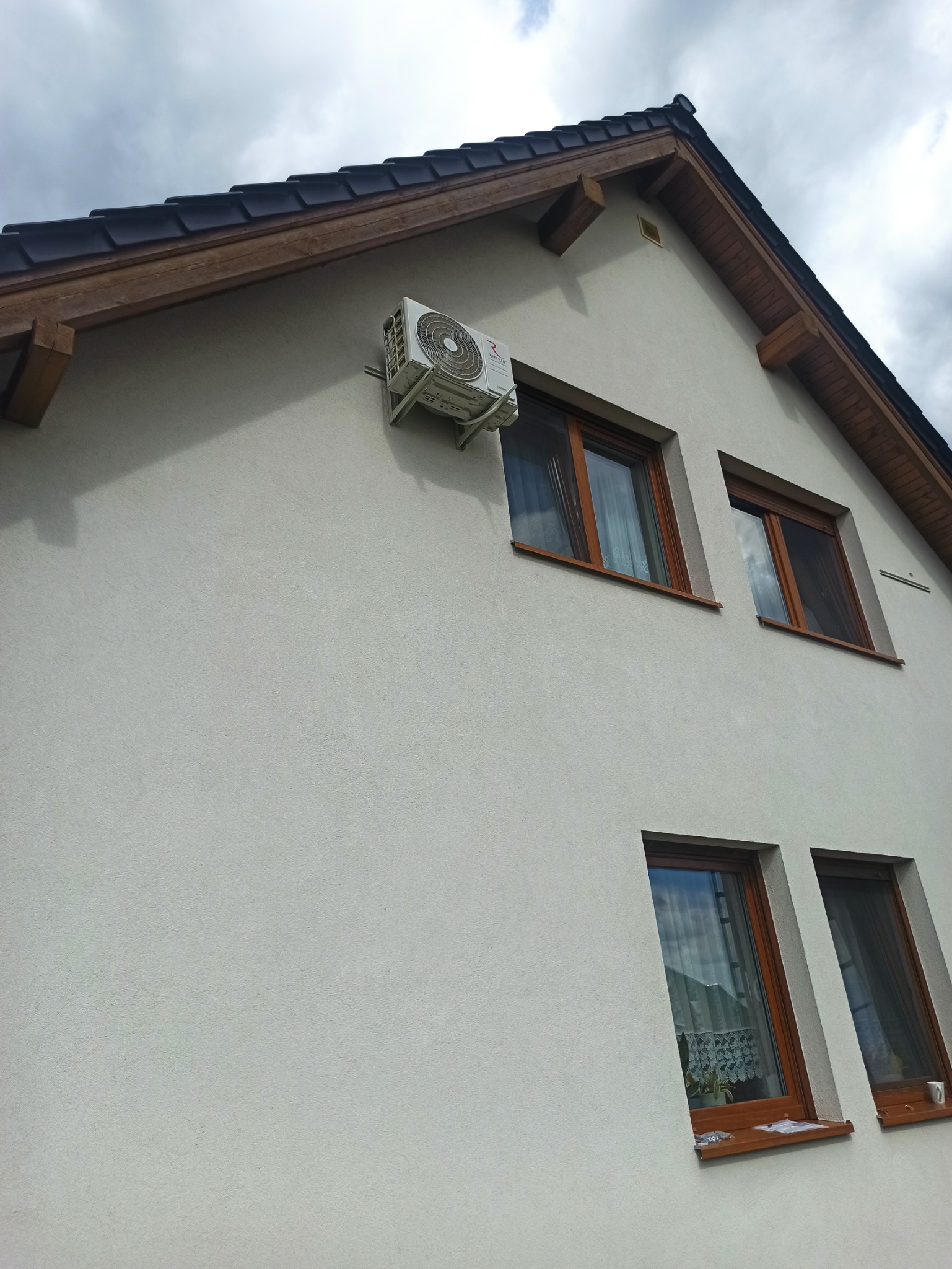 Biała elewacja domu z zamontowanym klimatyzatorem na wspornikach, widoczne okna z brązowymi ramami i fragment dachu pokrytego ciemną dachówką.