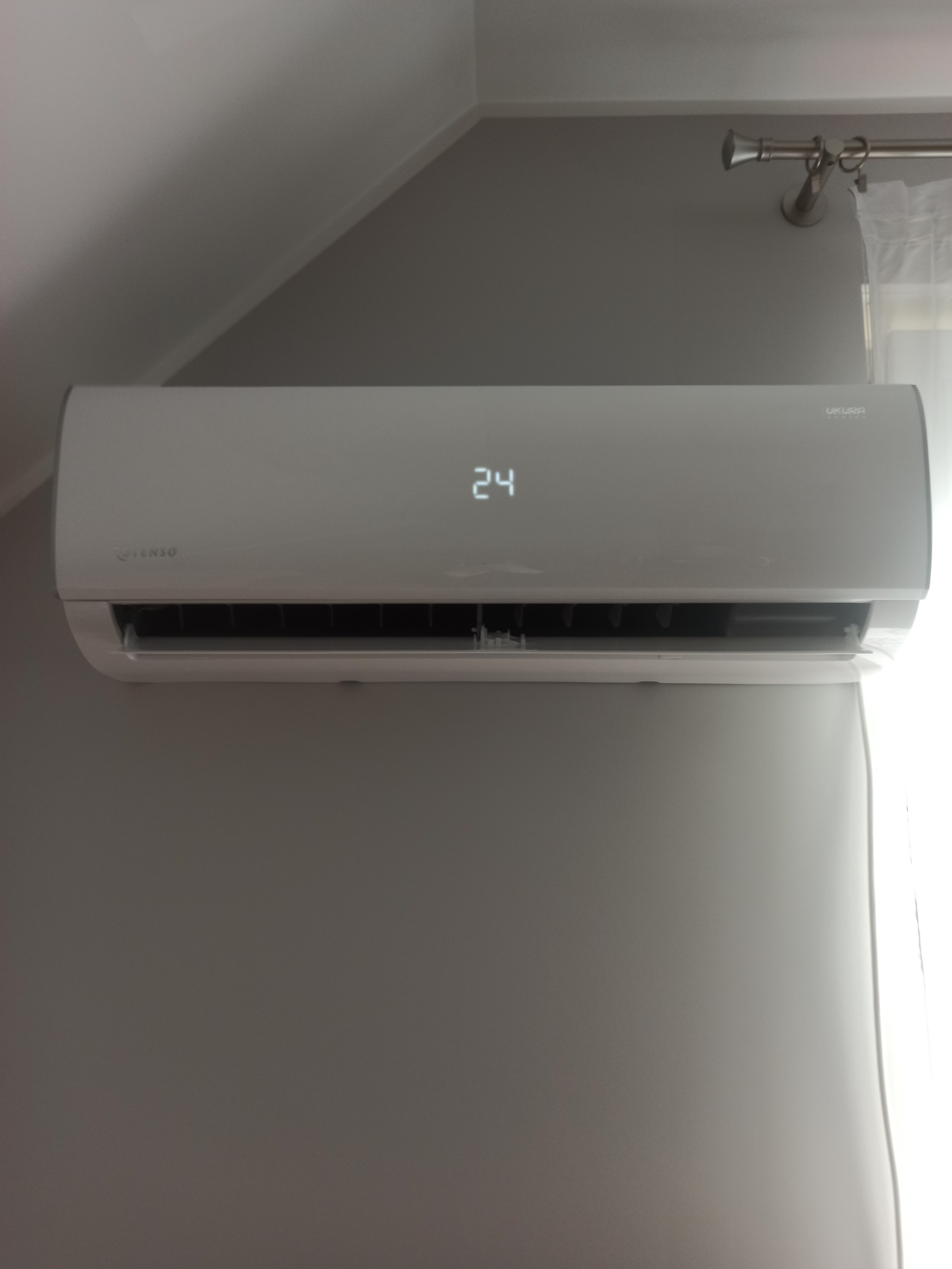 Biała klimatyzacja ścienna z wyświetlaną temperaturą 24 stopnie, zamontowana w szarym pokoju z karniszem i zasłoną w tle.