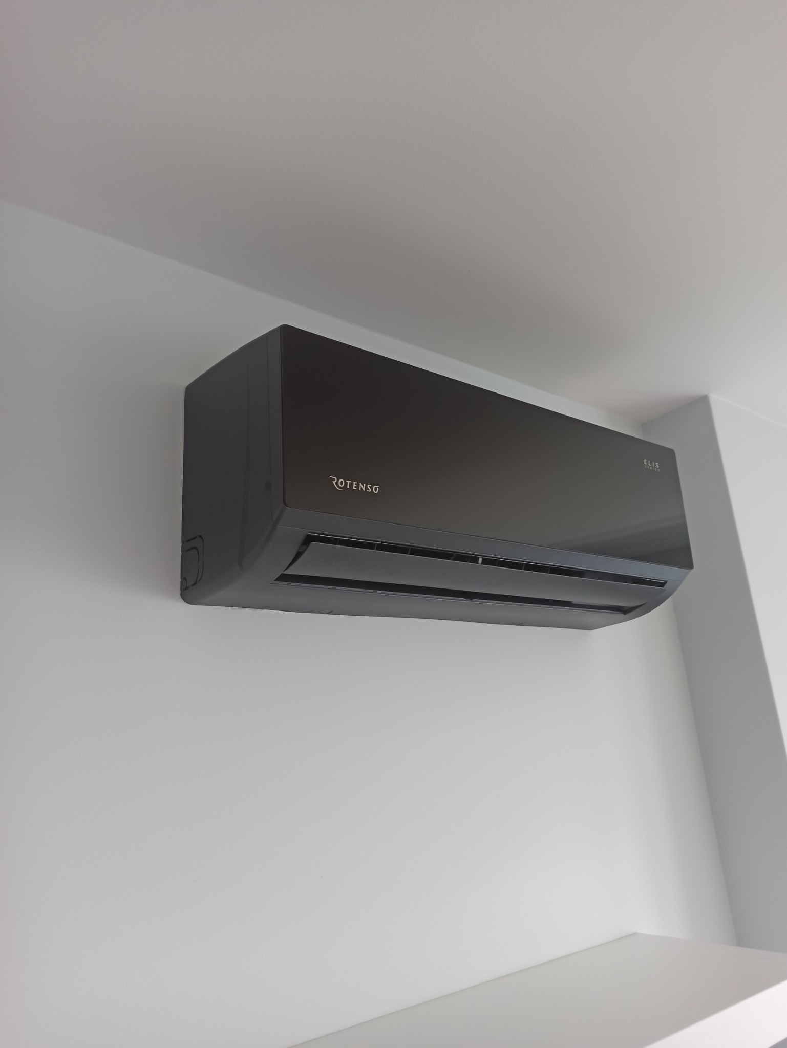 Czarny klimatyzator Rotenso ELIS zamontowany na białej ścianie w nowoczesnym wnętrzu. Widoczny z boku, z delikatnym oświetleniem.