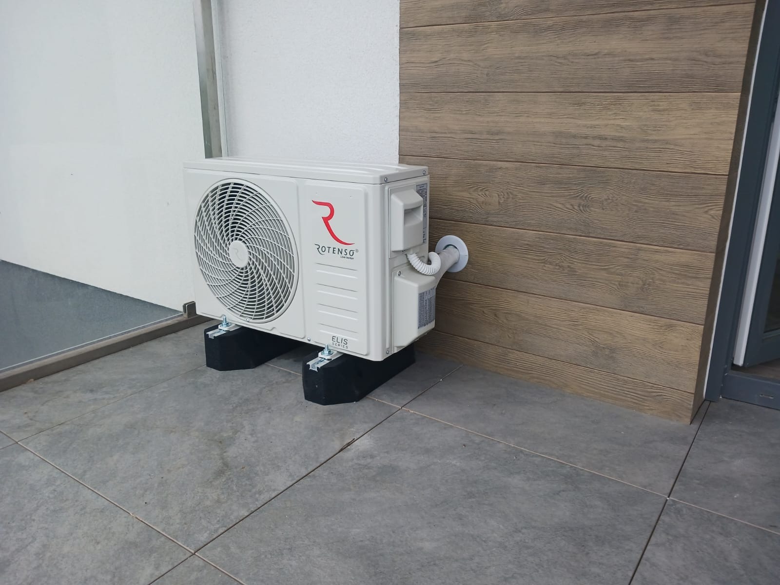 Zewnętrzna jednostka klimatyzacji Rotenso Elis Series, zamontowana na czarnych podkładkach antywibracyjnych, przy ścianie z drewnianą okładziną. Widoczny przewód odprowadzający skropliny.