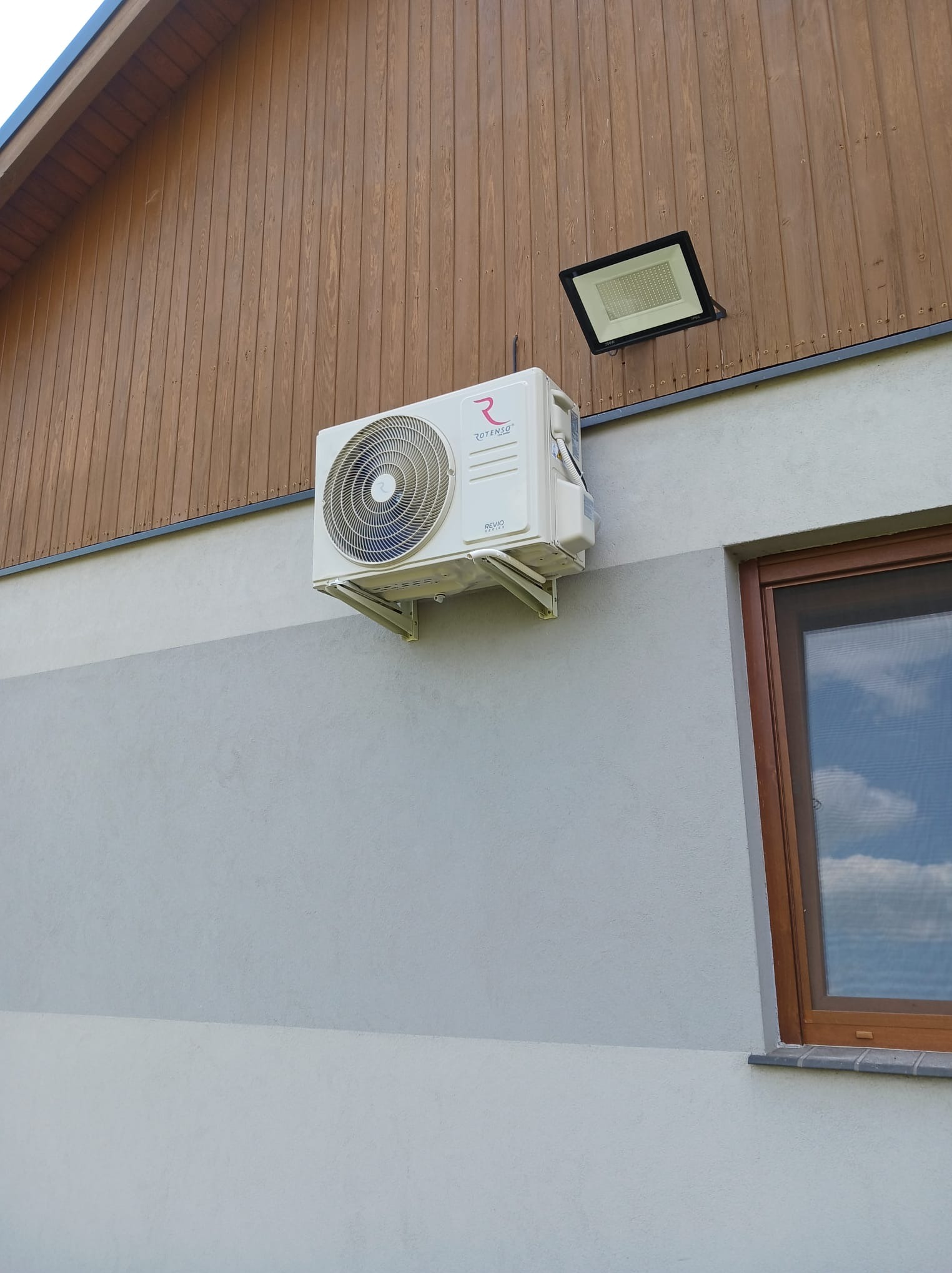 Biała jednostka zewnętrzna klimatyzacji Rotenso zamontowana na elewacji budynku z drewnianą boazerią i oknem. Widoczny reflektor oświetleniowy.