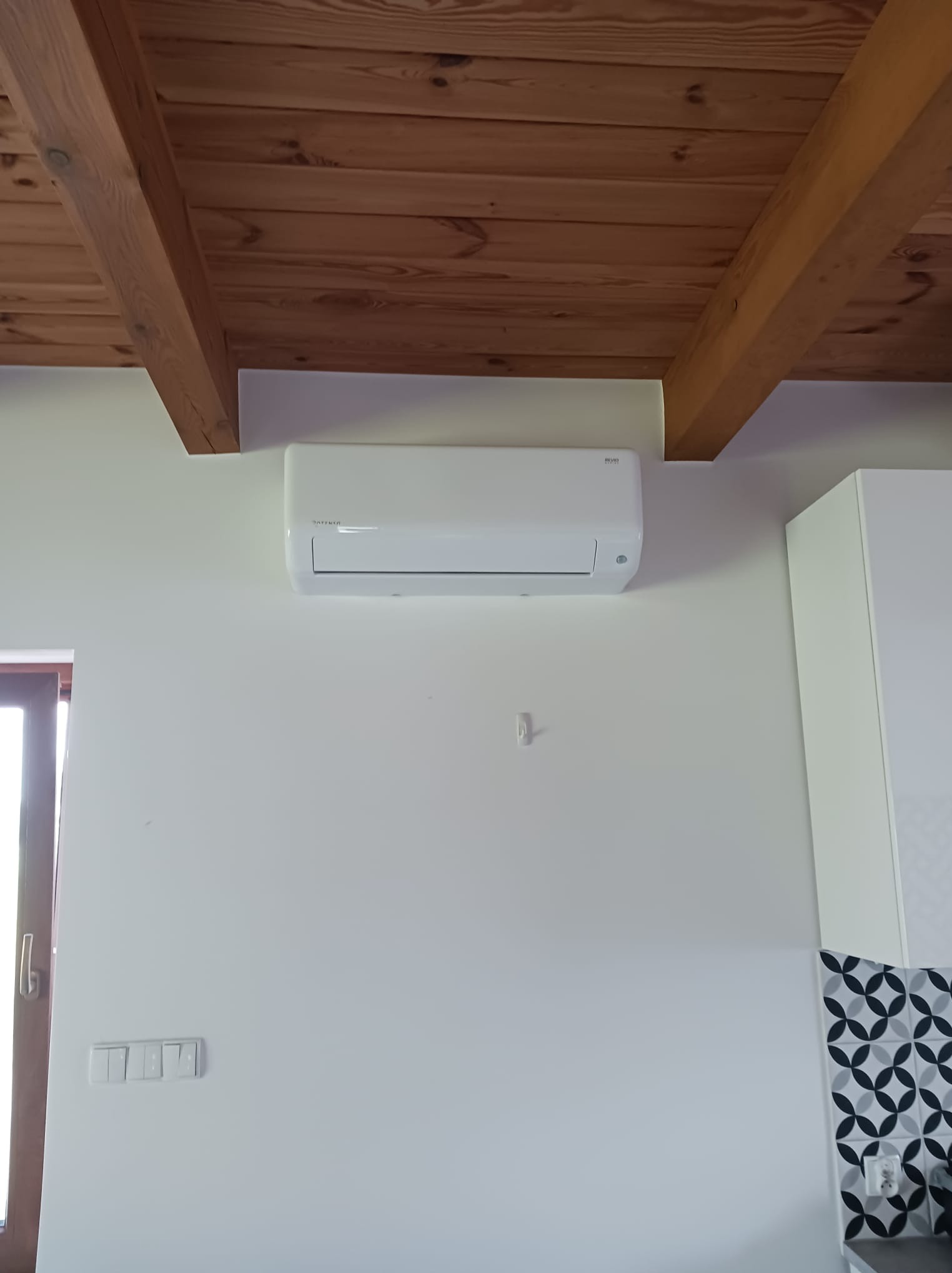 Biała klimatyzacja na ścianie w pomieszczeniu z drewnianym sufitem i fragmentem zabudowy kuchennej z dekoracyjnymi płytkami. Widoczne gniazdka i włączniki.