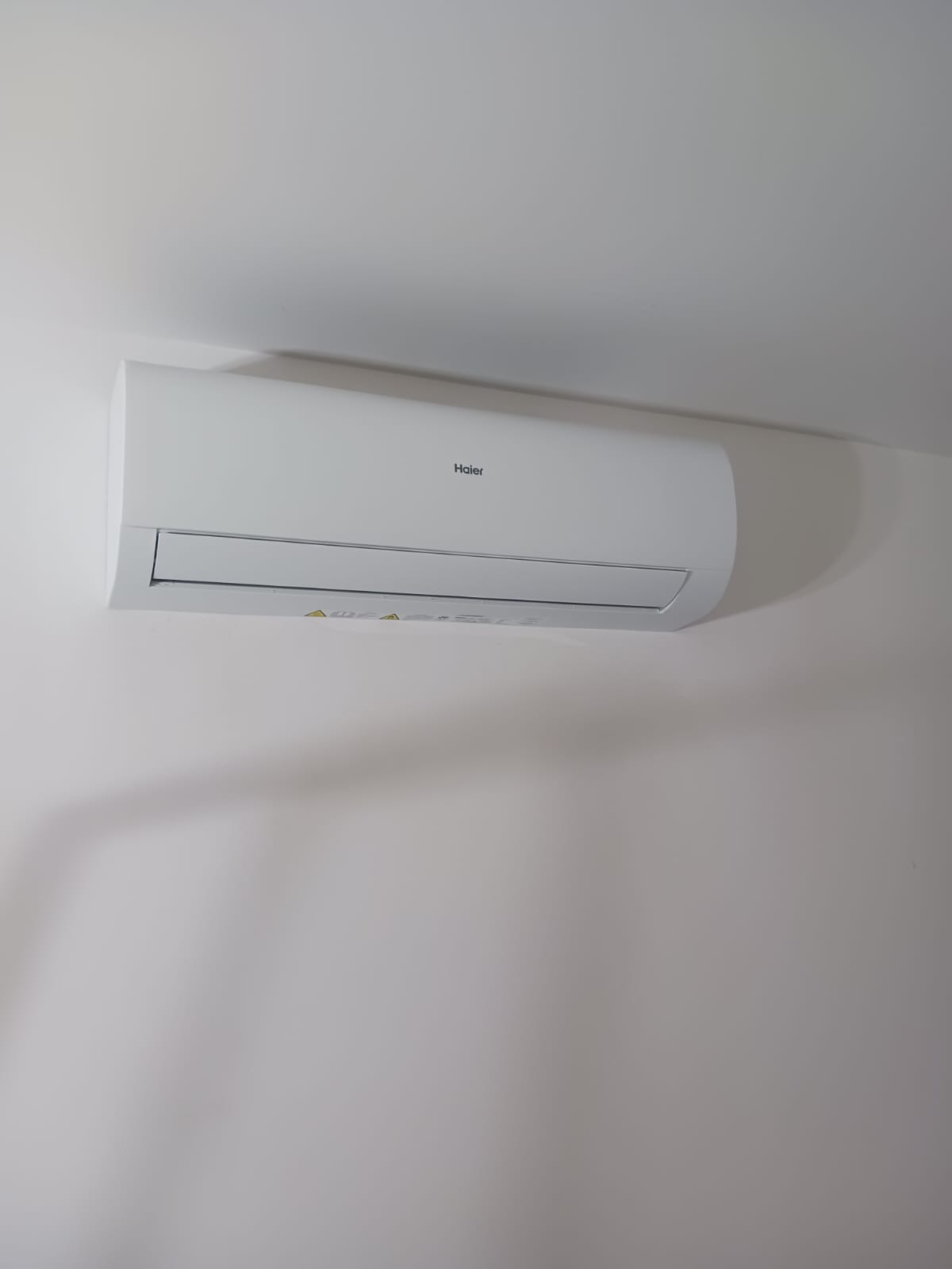 Biały klimatyzator Haier zamontowany na jasnej ścianie, widok z dołu, minimalistyczny design, oświetlenie dzienne.