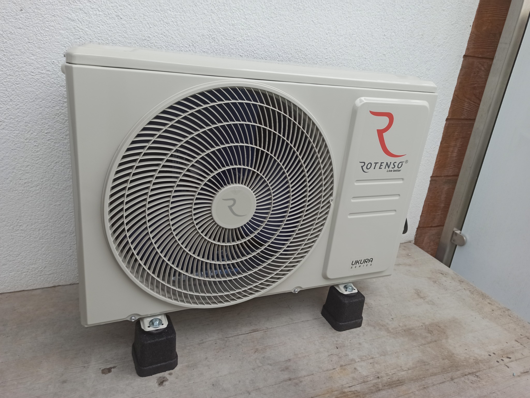 Biała jednostka zewnętrzna klimatyzacji Rotenso Ukura Series na czarnych podkładkach, ustawiona na drewnianej powierzchni przy białej ścianie. Widoczne logo i wentylator.