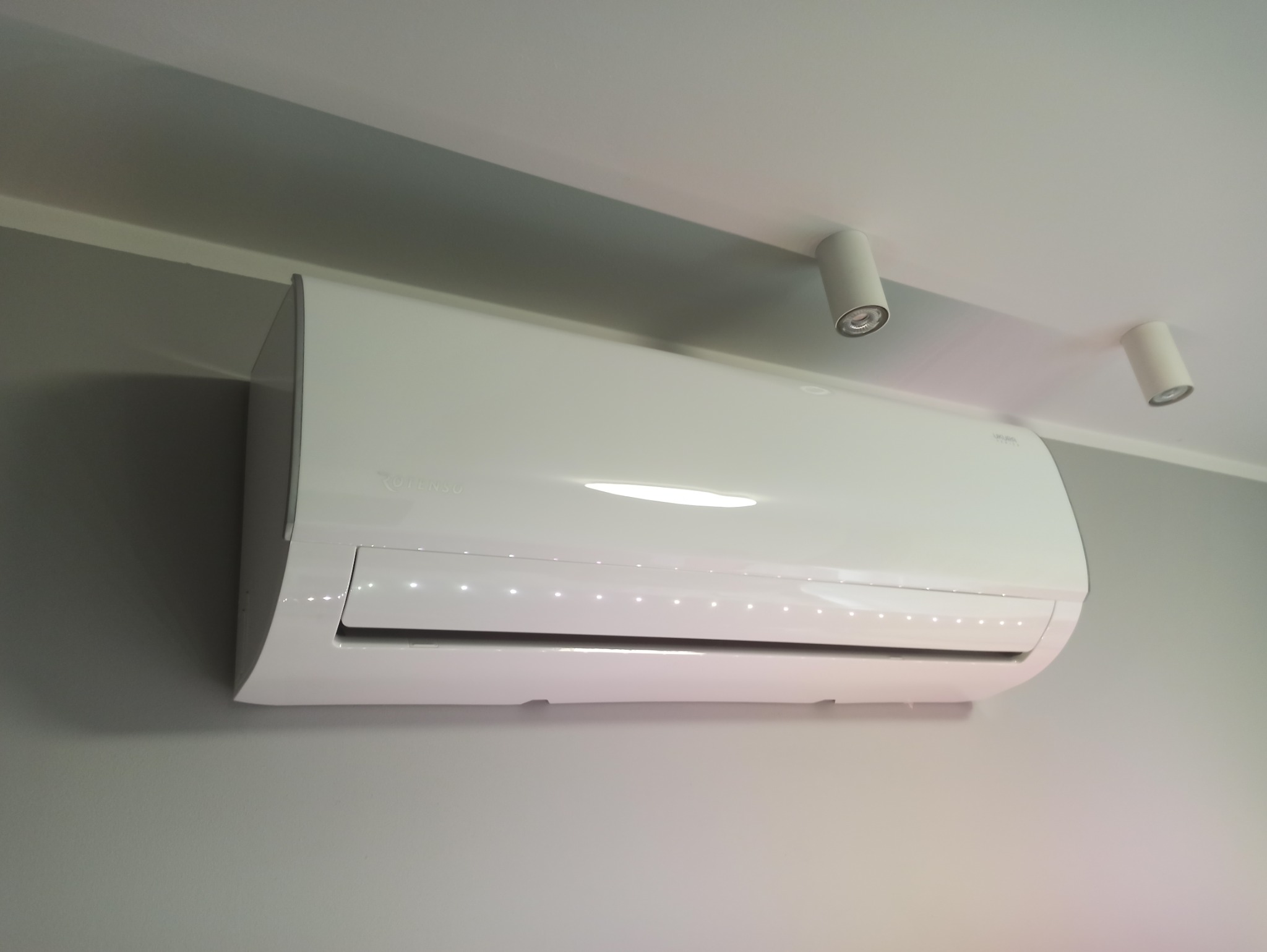 Biały klimatyzator Rotenso zamontowany na szarej ścianie, widoczne dwa białe reflektory sufitowe. Minimalistyczny design, nowoczesne oświetlenie.