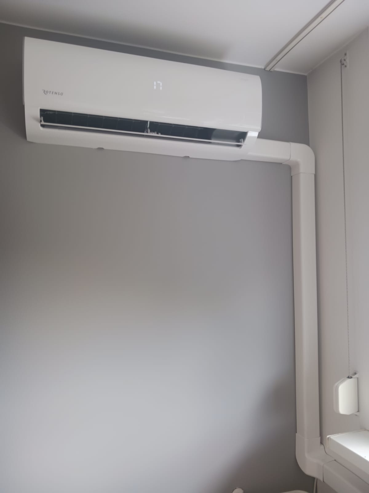 Biały klimatyzator Rotenso z widoczną rurą odprowadzającą, zamontowany na szarej ścianie. Na wyświetlaczu urządzenia widoczna temperatura 17 stopni.