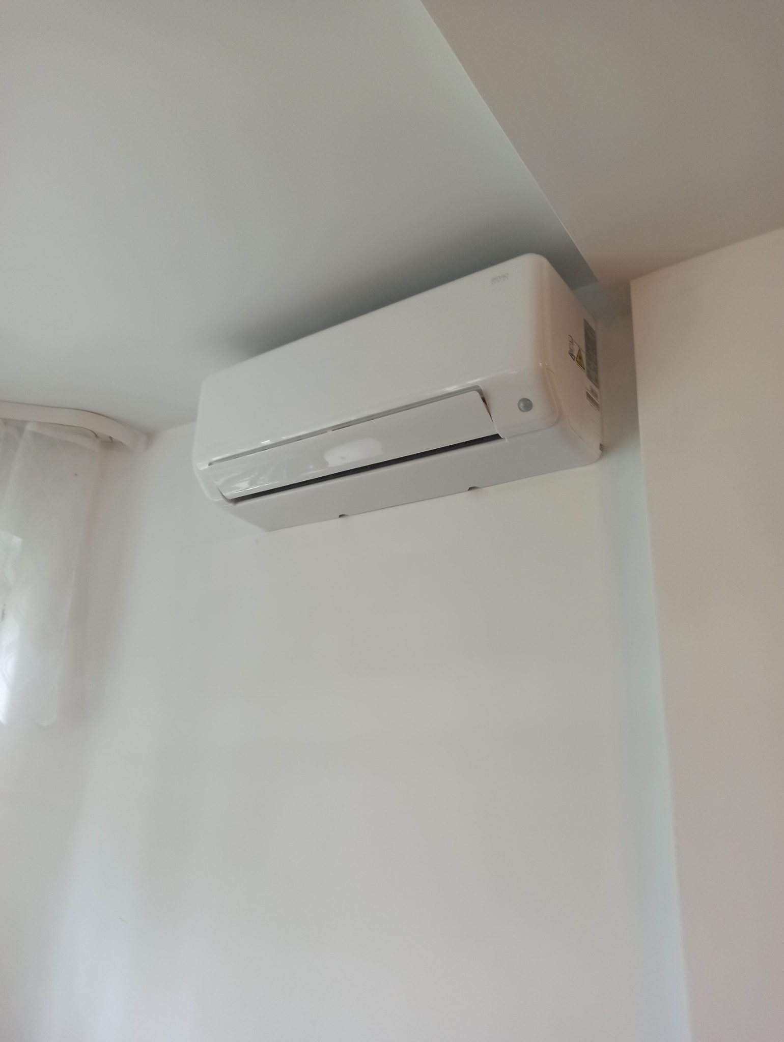 Biała klimatyzacja ścienna z widoczną folią ochronną na klapie, zamontowana w jasnym pomieszczeniu z oknem zasłoniętym firanką.