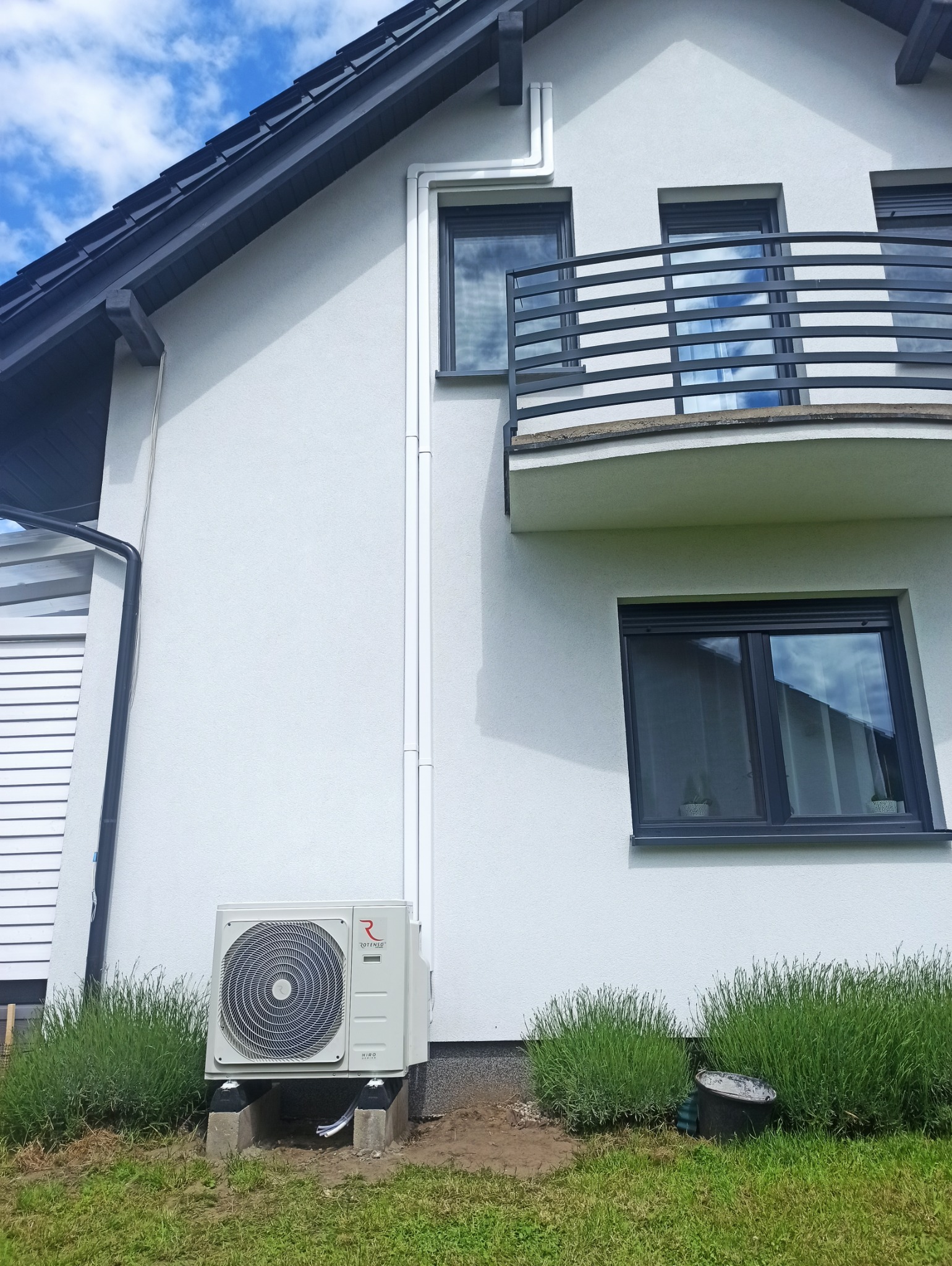 Biała jednostka zewnętrzna klimatyzacji Rotenso zamontowana przy ścianie domu z balkonem i szarą stolarką okienną. Widoczne elementy instalacji.