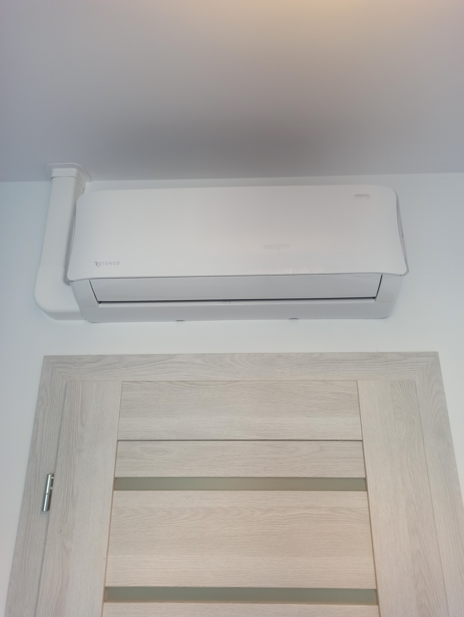 Biały klimatyzator Rotenso zamontowany nad drzwiami o jasnym, drewnopodobnym wykończeniu, widoczny na tle białej ściany.