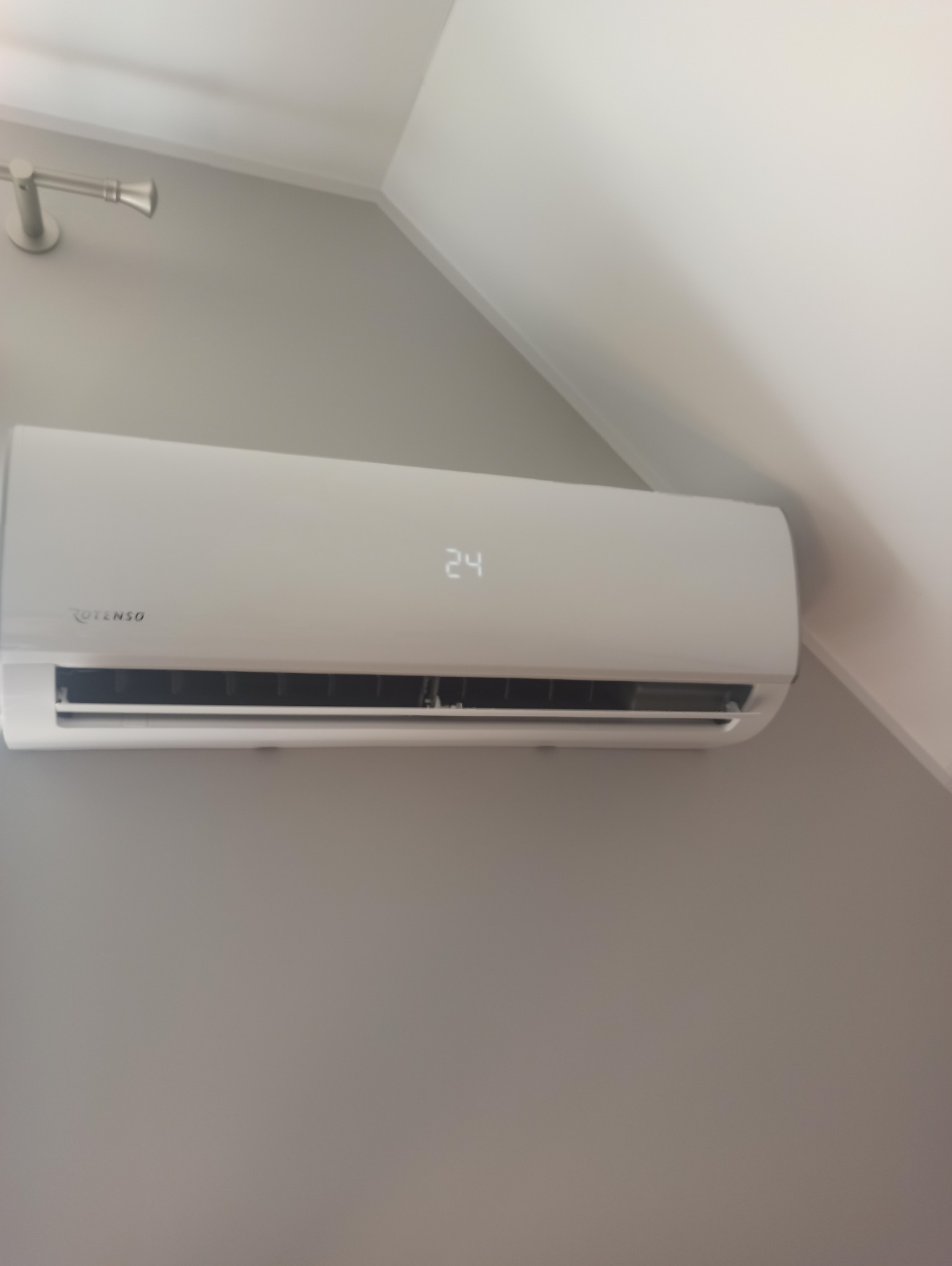 Biały klimatyzator Rotenso na szarej ścianie, ustawiony na 24 stopnie, widoczny fragment sufitu i ściany pod kątem.