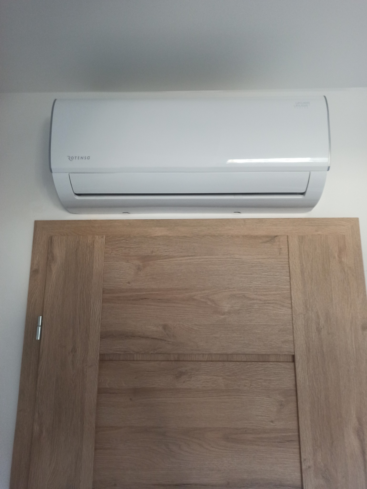 Biała klimatyzacja Rotenso, zamontowana nad drewnianymi drzwiami w jasnym wnętrzu. Widok z dołu, prezentacja standardowego montażu.