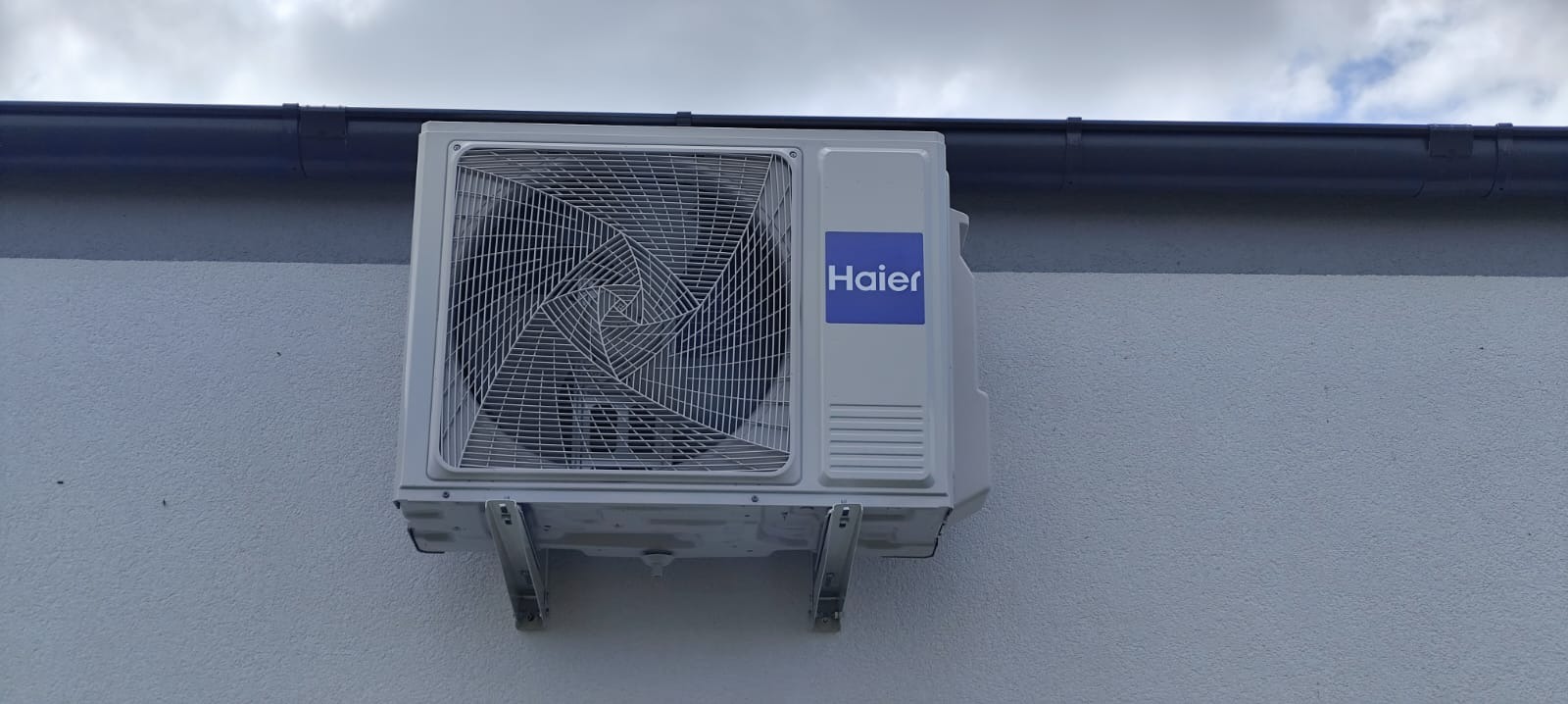 Zamontowana na zewnątrz jednostka klimatyzacji Haier na jasnej ścianie, widoczny fragment rynny i pochmurne niebo w tle. Kąt ujęcia od dołu.