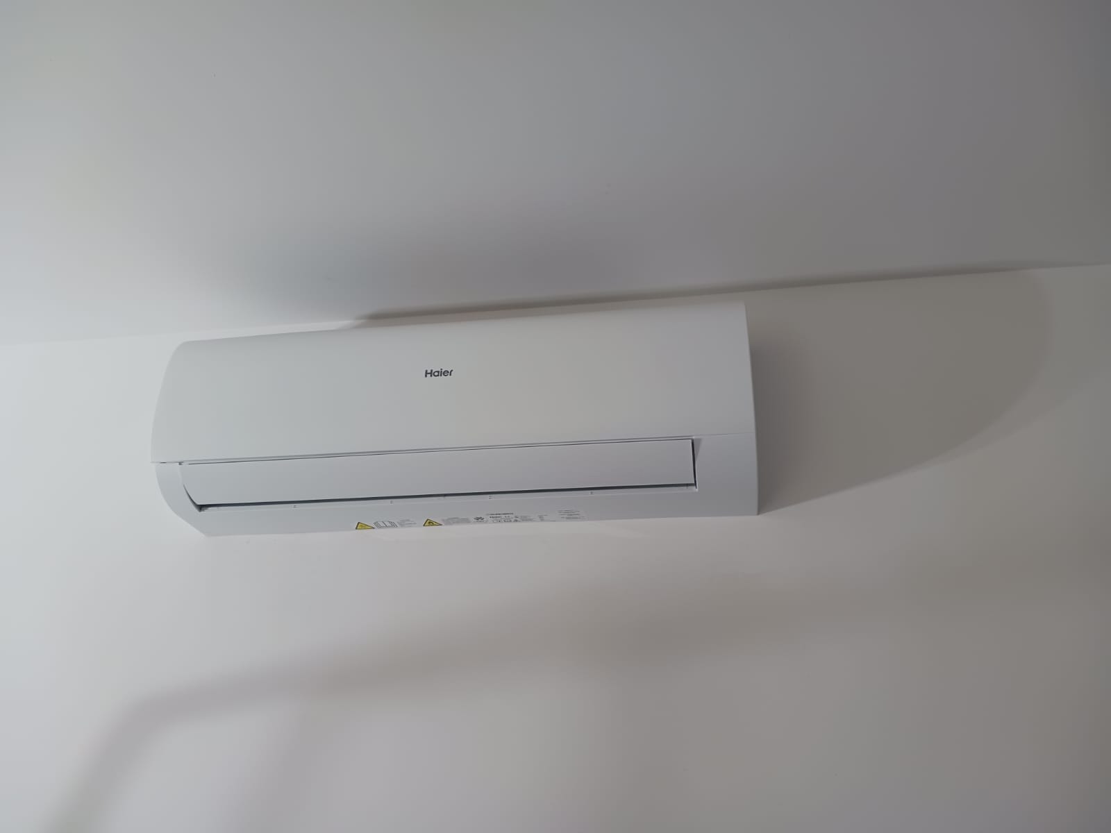 Biały klimatyzator Haier zamontowany na jasnej ścianie, z widocznymi detalami obudowy i subtelnym cieniem, sugerujący świeżą instalację w nowoczesnym wnętrzu.