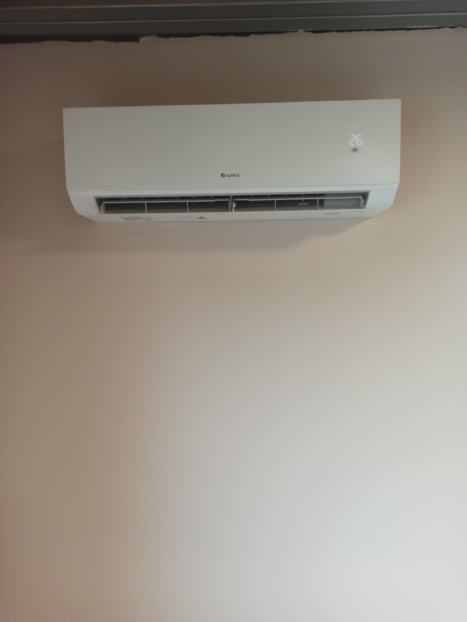 Biały klimatyzator Gree zamontowany na beżowej ścianie. Wyświetlacz pokazuje temperaturę 26 stopni. Widoczny fragment sufitu z szarą listwą.