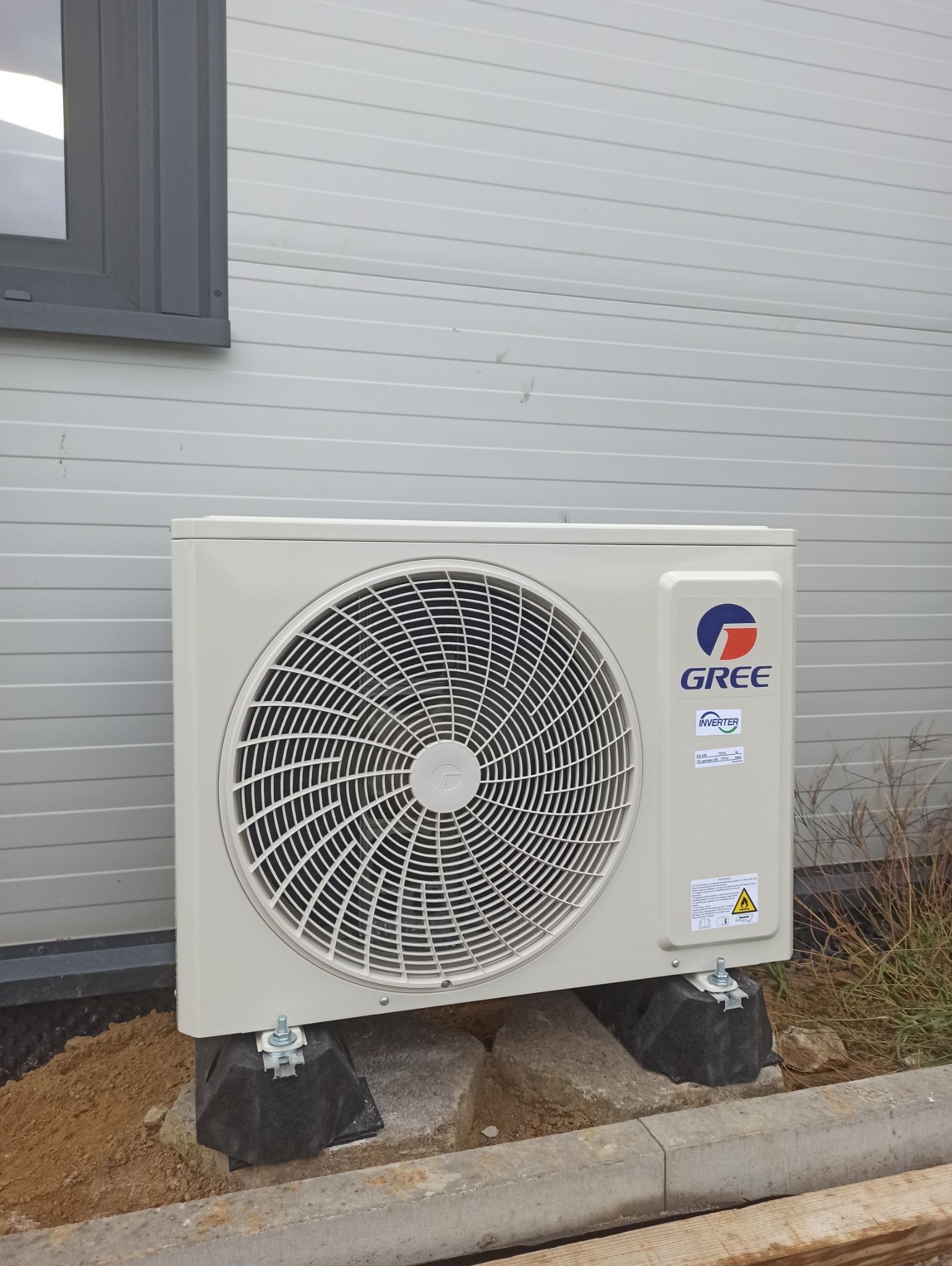 Zamontowana jednostka zewnętrzna klimatyzacji GREE na elewacji budynku z szarego sidingu, na betonowych bloczkach, widoczne fragmenty trawy i okno.