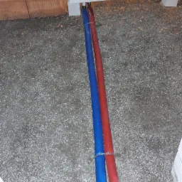 Podłączenie rur grzewczych: niebieska i czerwona, wychodzących spod białej skrzynki w ścianie z cegieł, ułożone na surowym betonowym podłożu, z widocznymi złączkami i opaskami zaciskowymi.