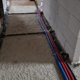 Instalacja ogrzewania podłogowego w trakcie montażu: widoczne rury grzewcze (niebieska i czerwona) oraz czarne rury elektryczne mocowane do surowej posadzki w nowym budynku z bloczków.