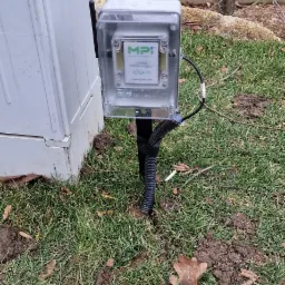 Zewnętrzny czujnik poziomu cieczy Aquato MPI zamontowany na metalowym słupku, z kablem w peszlu, obok białej skrzynki na trawniku.