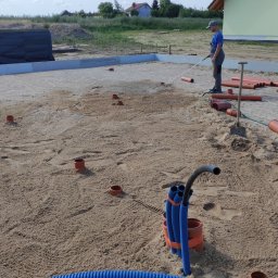 Halina Alsanowska - Przygotowanie terenu pod fundamenty domu: ubijanie piasku wodą, widoczne rury kanalizacyjne i odwodnieniowe, wykopane otwory pod słupki.