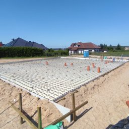 Halina Alsanowska - Przygotowany zbrojony fundament pod budowę domu jednorodzinnego, widoczne rury kanalizacyjne wystające pionowo, szalunki i piasek.