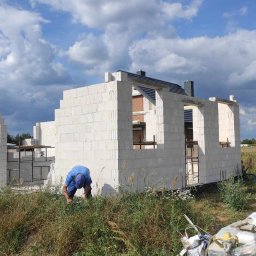 Halina Alsanowska - Wznoszenie ścian domu z białych bloczków, widoczny fragment dachu, pracownik w niebieskim ubraniu przy murze, trawa i nieporządek na pierwszym planie.