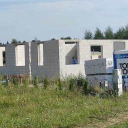 Halina Alsanowska - Surowy stan budynku z betonu komórkowego z widocznym pracownikiem w niebieskiej koszulce, sterta materiałów budowlanych i przenośna toaleta Toi Toi na zarośniętej działce.