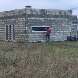 Budowa domu murowanego: murarz w czerwonej koszuli montuje okno w surowej ścianie z bloczków, widoczne worki z zaprawą i niezagospodarowany teren wokół.