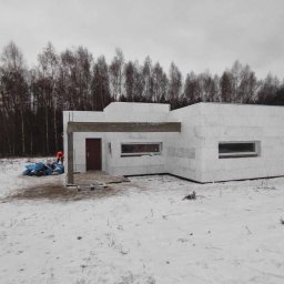 Halina Alsanowska - Surowy stan nowoczesnego domu z białych bloczków w zimowym krajobrazie, widoczne okna i drewniany element konstrukcyjny nad wejściem, pracownik w pomarańczowej kamizelce w tle.