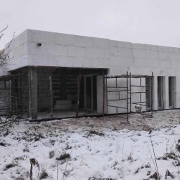 Halina Alsanowska - Budowa domu jednorodzinnego w zimowej scenerii; ściany wzniesione z białych bloczków, widoczne rusztowania i przygotowane otwory okienne.