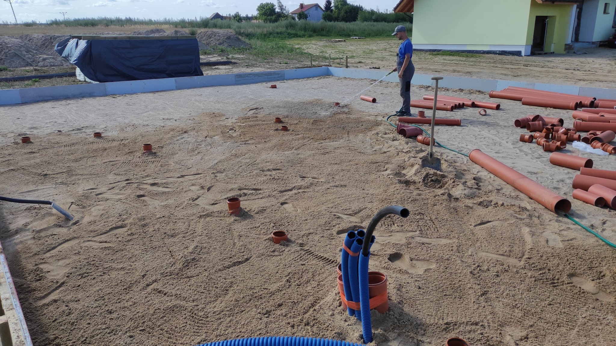 Przygotowanie terenu pod fundamenty domu: ubijanie piasku wodą, widoczne rury kanalizacyjne i odwodnieniowe, wykopane otwory pod słupki.